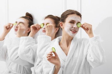 Photo of Happy women with facial masks and cucumber slices in bathrobes after spa procedures near white wall, selective focus