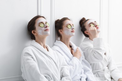 Photo of Happy women with facial masks and cucumber slices in bathrobes after spa procedures near white wall
