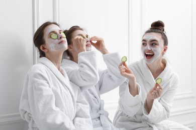 Photo of Happy women with facial masks and cucumber slices in bathrobes after spa procedures near white wall