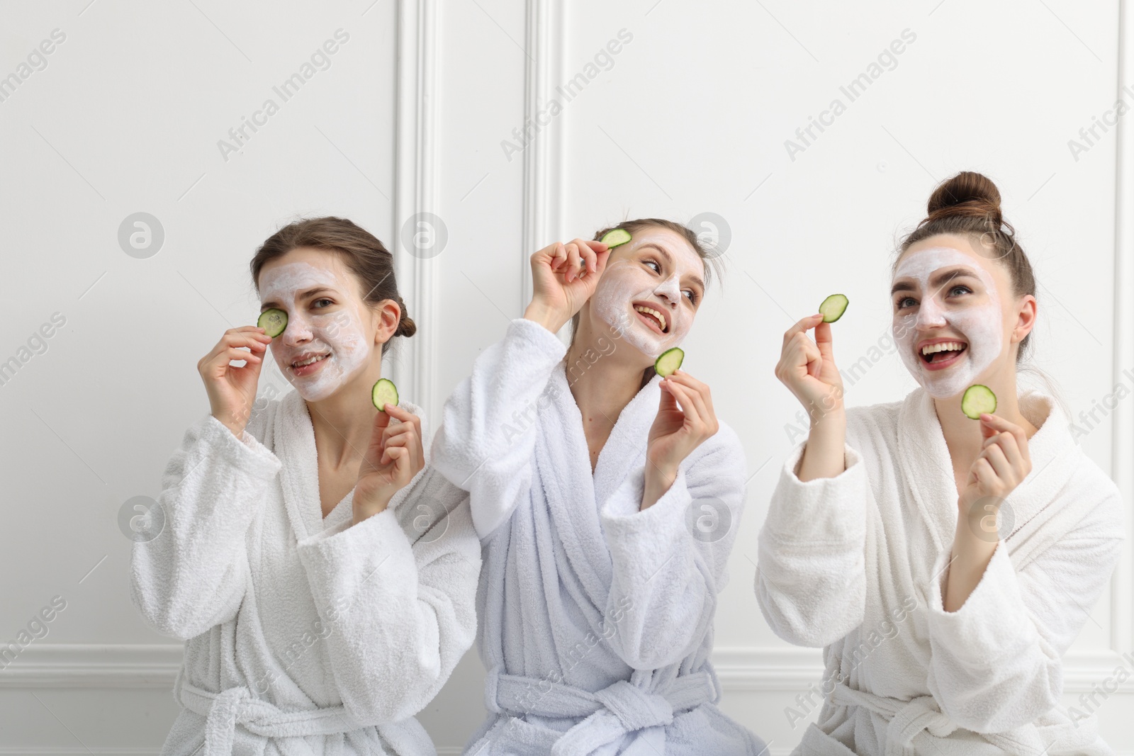 Photo of Happy women with facial masks and cucumber slices in bathrobes after spa procedures near white wall