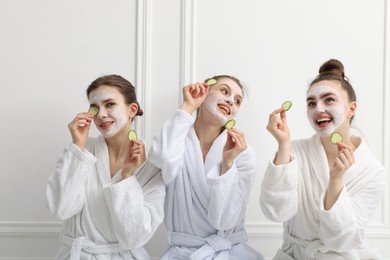 Photo of Happy women with facial masks and cucumber slices in bathrobes after spa procedures near white wall