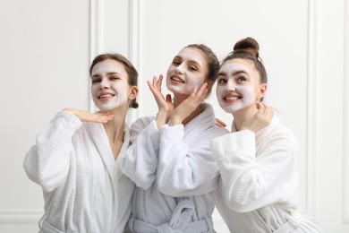 Photo of Happy women with facial masks in bathrobes posing after spa procedures near white wall