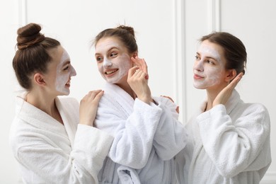 Photo of Happy women with facial masks in bathrobes posing after spa procedures near white wall