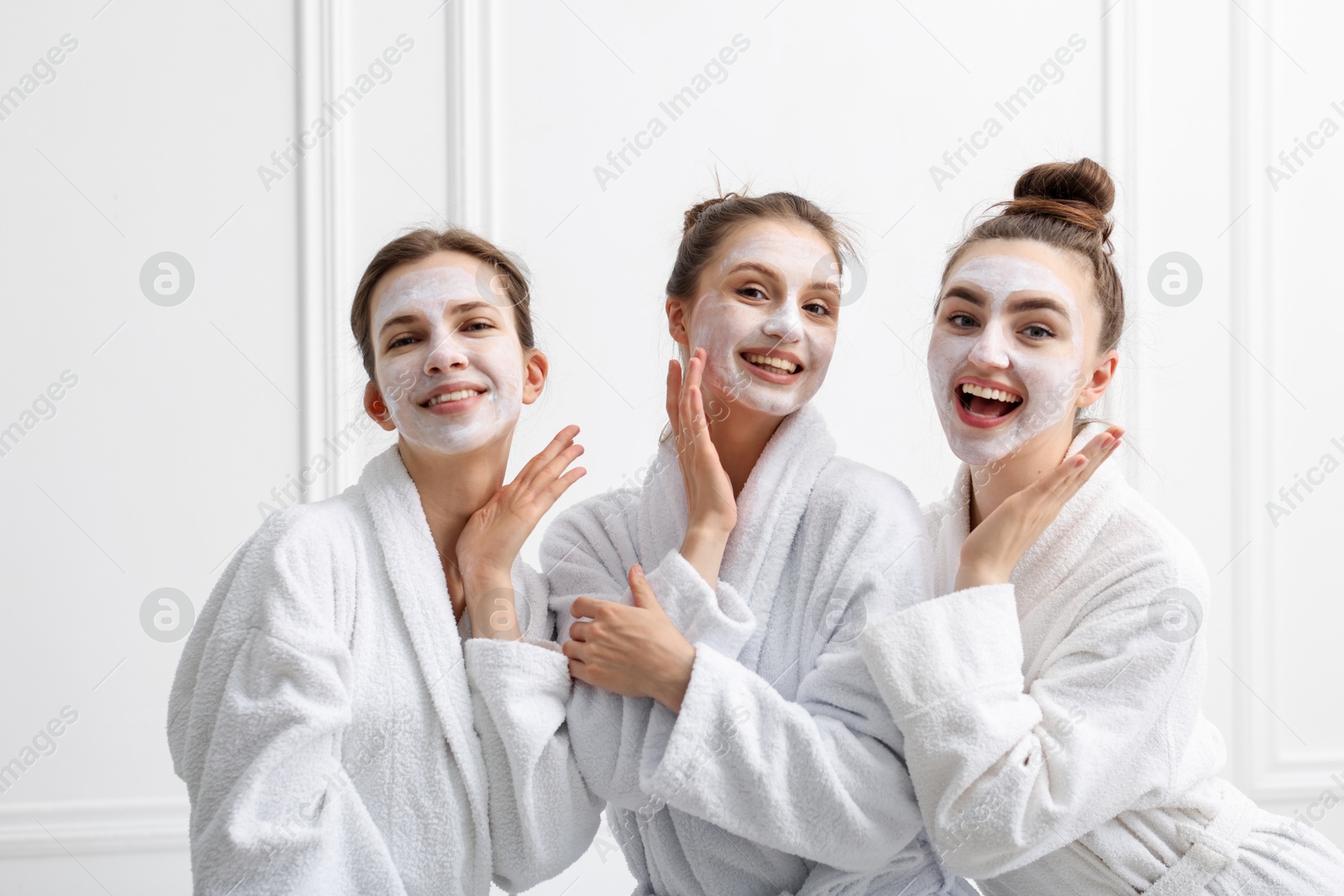 Photo of Happy women with facial masks in bathrobes posing after spa procedures near white wall