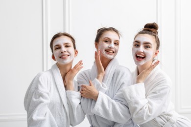 Photo of Happy women with facial masks in bathrobes posing after spa procedures near white wall