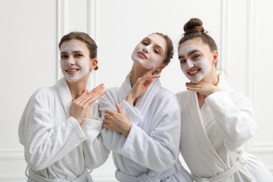 Photo of Happy women with facial masks in bathrobes posing after spa procedures near white wall