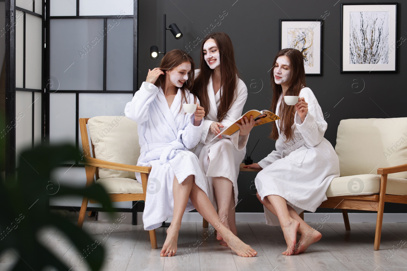 Photo of Happy women with facial masks and cups of healthy drink reading magazine in bathrobes after spa procedures indoors