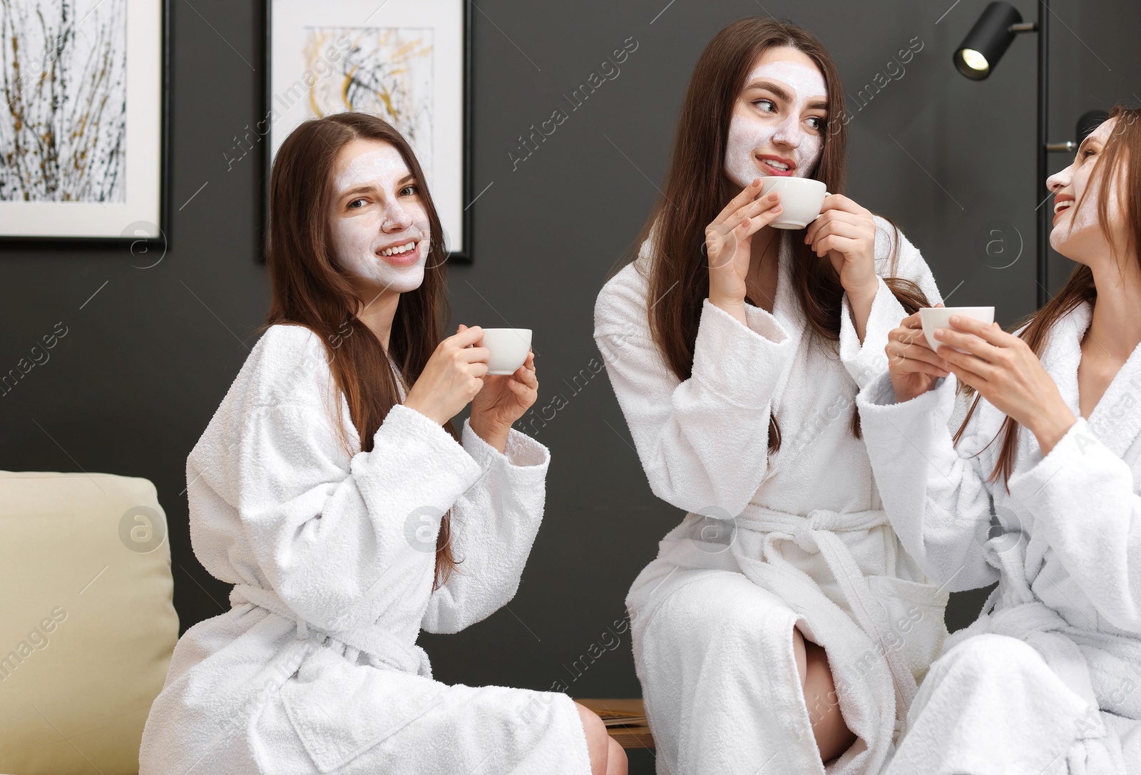 Photo of Happy women with facial masks and cups of healthy drink in bathrobes after spa procedures indoors