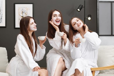 Photo of Happy women with facial masks and cups of healthy drink in bathrobes after spa procedures indoors