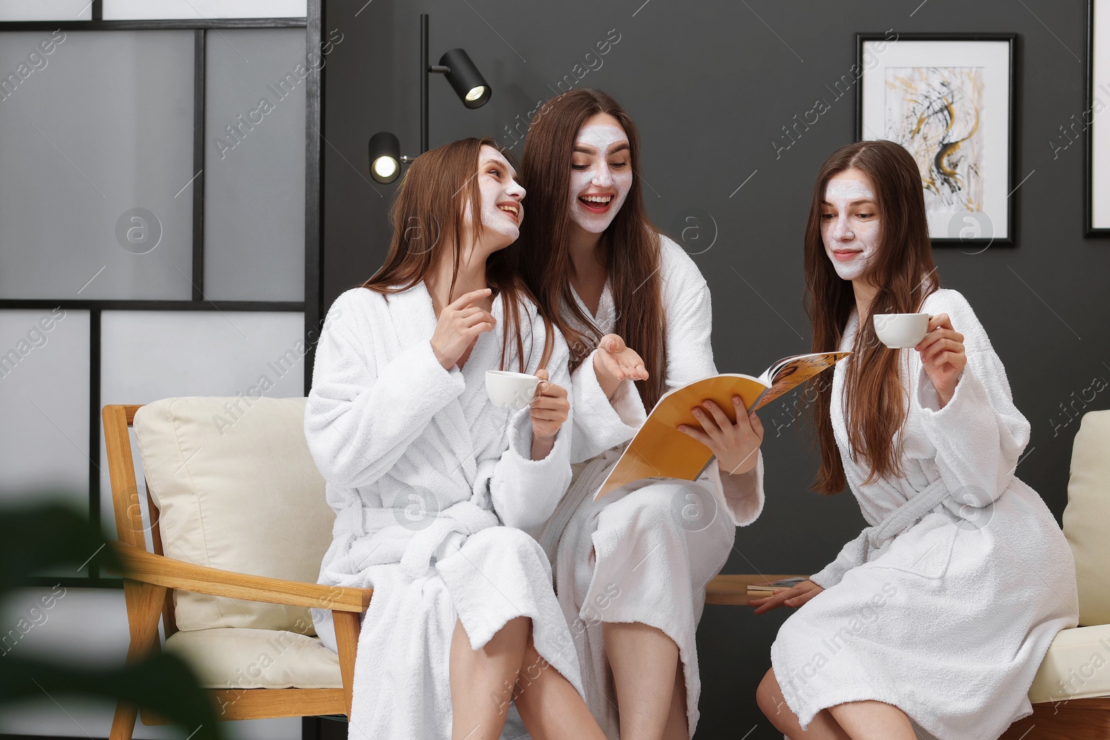 Photo of Happy women with facial masks and cups of healthy drink reading magazine in bathrobes after spa procedures indoors