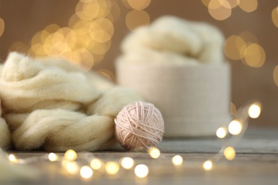 Photo of Soft felting wool and ball of yarn on wooden table against brown background with blurred lights, closeup