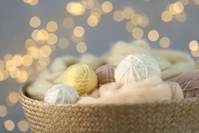 Photo of Soft balls of yarn and felting wool in wicker basket against grey background with blurred lights, closeup