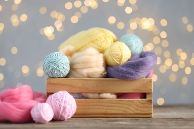 Photo of Soft balls of yarn and felting wool in crate on wooden table against grey background with blurred lights, closeup