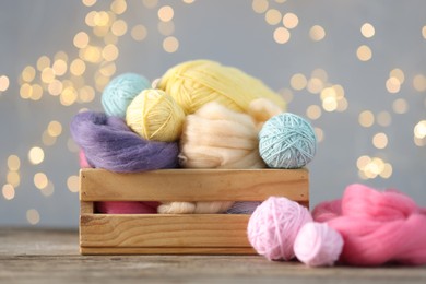 Photo of Soft balls of yarn and felting wool in crate on wooden table against grey background with blurred lights, closeup