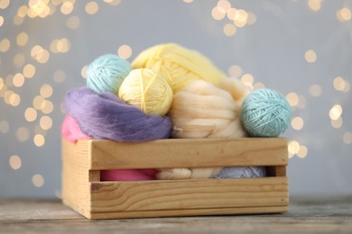 Photo of Soft balls of yarn and felting wool in crate on wooden table against grey background with blurred lights, closeup