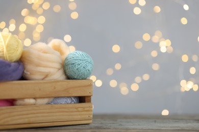 Photo of Soft balls of yarn and felting wool in crate on wooden table against grey background with blurred lights, closeup. Space for text