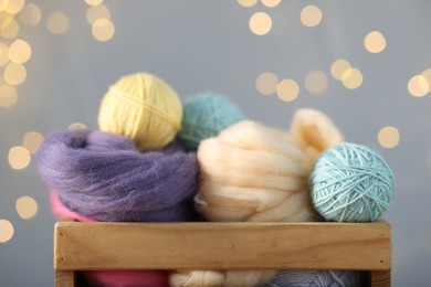 Photo of Soft balls of yarn and felting wool in crate against grey background with blurred lights, closeup