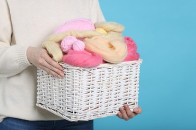 Photo of Woman holding wicker basket with colorful felting wool and yarns on light blue background, closeup