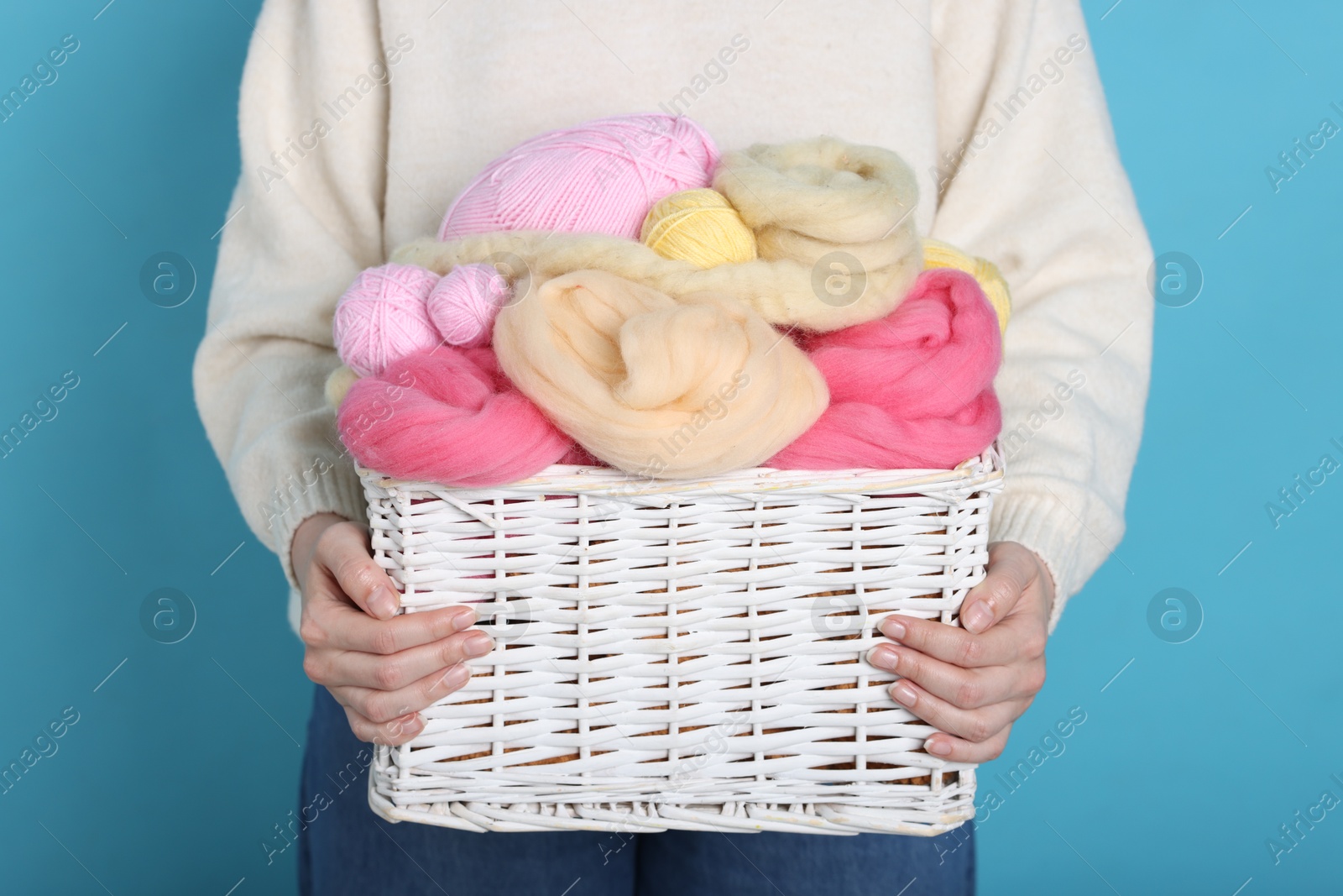 Photo of Woman holding wicker basket with colorful felting wool and yarns on light blue background, closeup