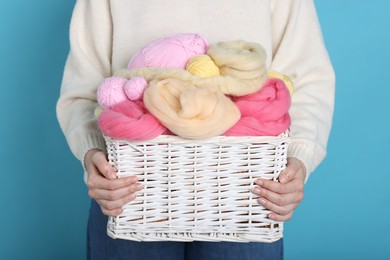 Photo of Woman holding wicker basket with colorful felting wool and yarns on light blue background, closeup