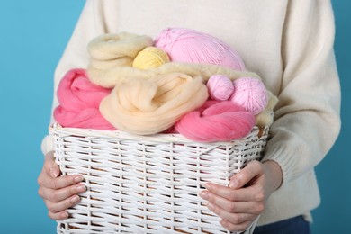 Photo of Woman holding wicker basket with colorful felting wool and yarns on light blue background, closeup