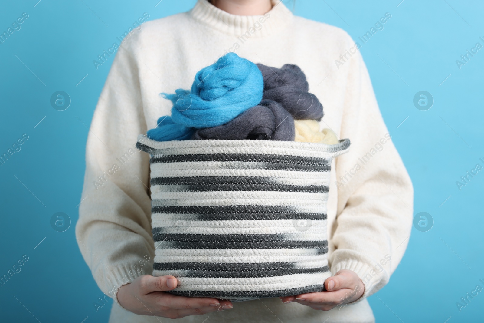 Photo of Woman holding wicker basket with colorful felting wool on light blue background, closeup