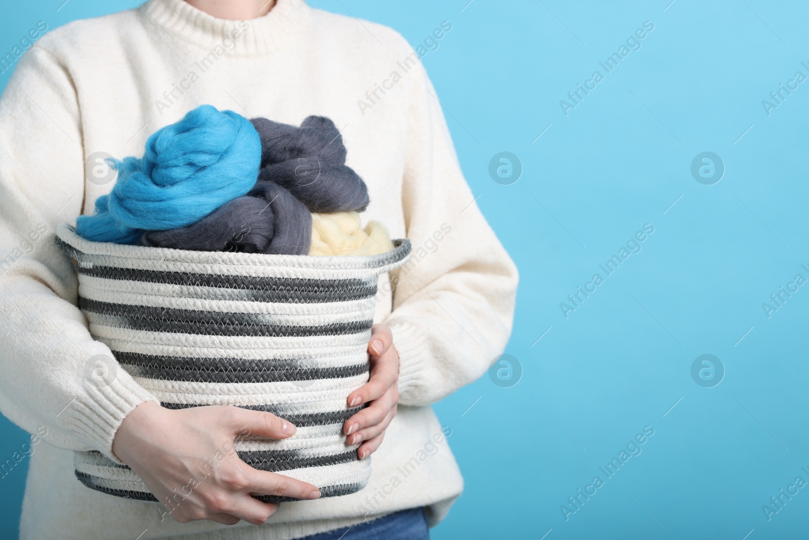 Photo of Woman holding wicker basket with colorful felting wool on light blue background, closeup. Space for text