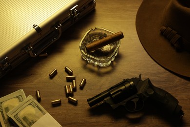 Photo of Mafia and criminal world. Gun, bullets, ashtray with cigar, hat, money and briefcase on wooden table, closeup