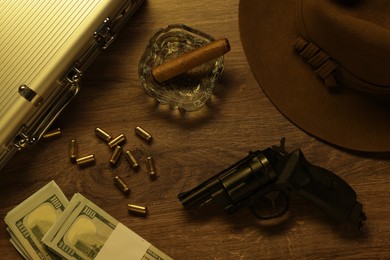 Photo of Mafia and criminal world. Gun, bullets, ashtray with cigar, hat, money and briefcase on wooden table, flat lay