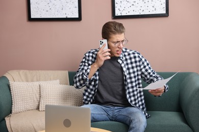 Photo of Young man with laptop and papers complaining to someone via phone at home