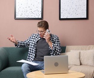 Photo of Young man with laptop and papers complaining to someone via phone at home