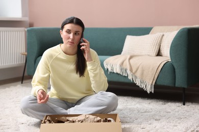 Photo of Young woman with package complaining to customer service via phone at home