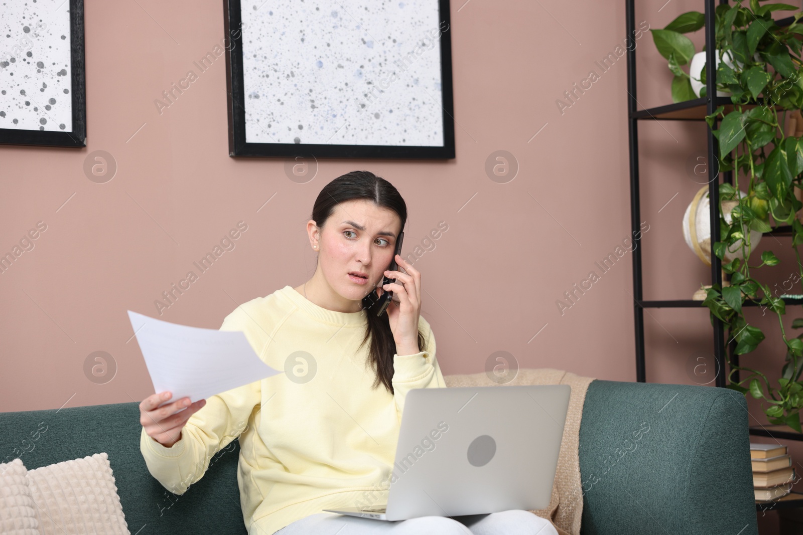 Photo of Young woman with laptop and papers complaining to someone via phone at home