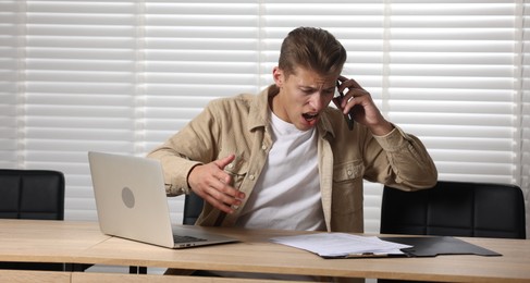 Photo of Young man with papers and laptop complaining to someone via phone at wooden table indoors