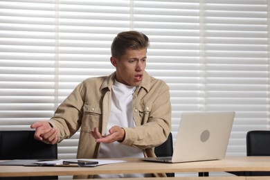 Photo of Young man with papers complaining to someone via laptop at wooden table indoors