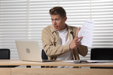 Photo of Young man with papers and laptop complaining to someone via phone at wooden table indoors