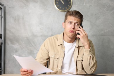 Photo of Young man with papers complaining to someone via phone at wooden table indoors