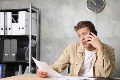 Photo of Young man with papers complaining to someone via phone at wooden table indoors