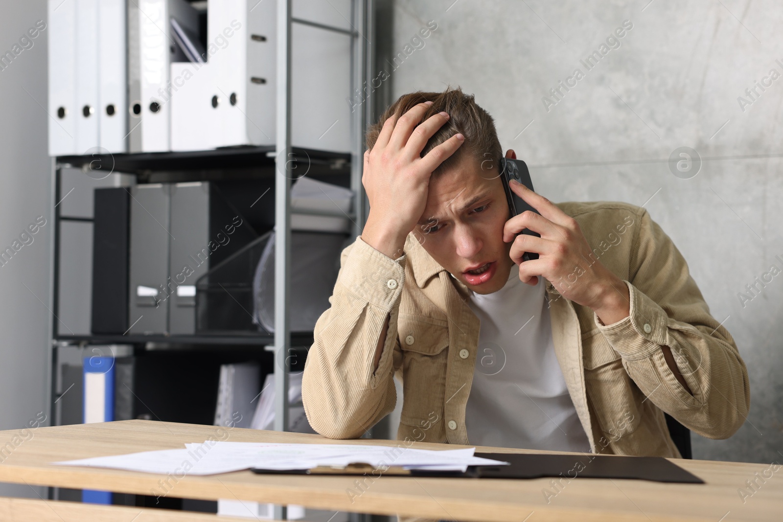 Photo of Young man with papers complaining to someone via phone at wooden table indoors
