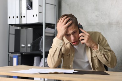 Photo of Young man with papers complaining to someone via phone at wooden table indoors