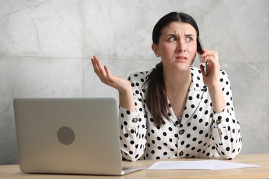 Photo of Young businesswoman with papers and laptop complaining to someone via phone at wooden table indoors