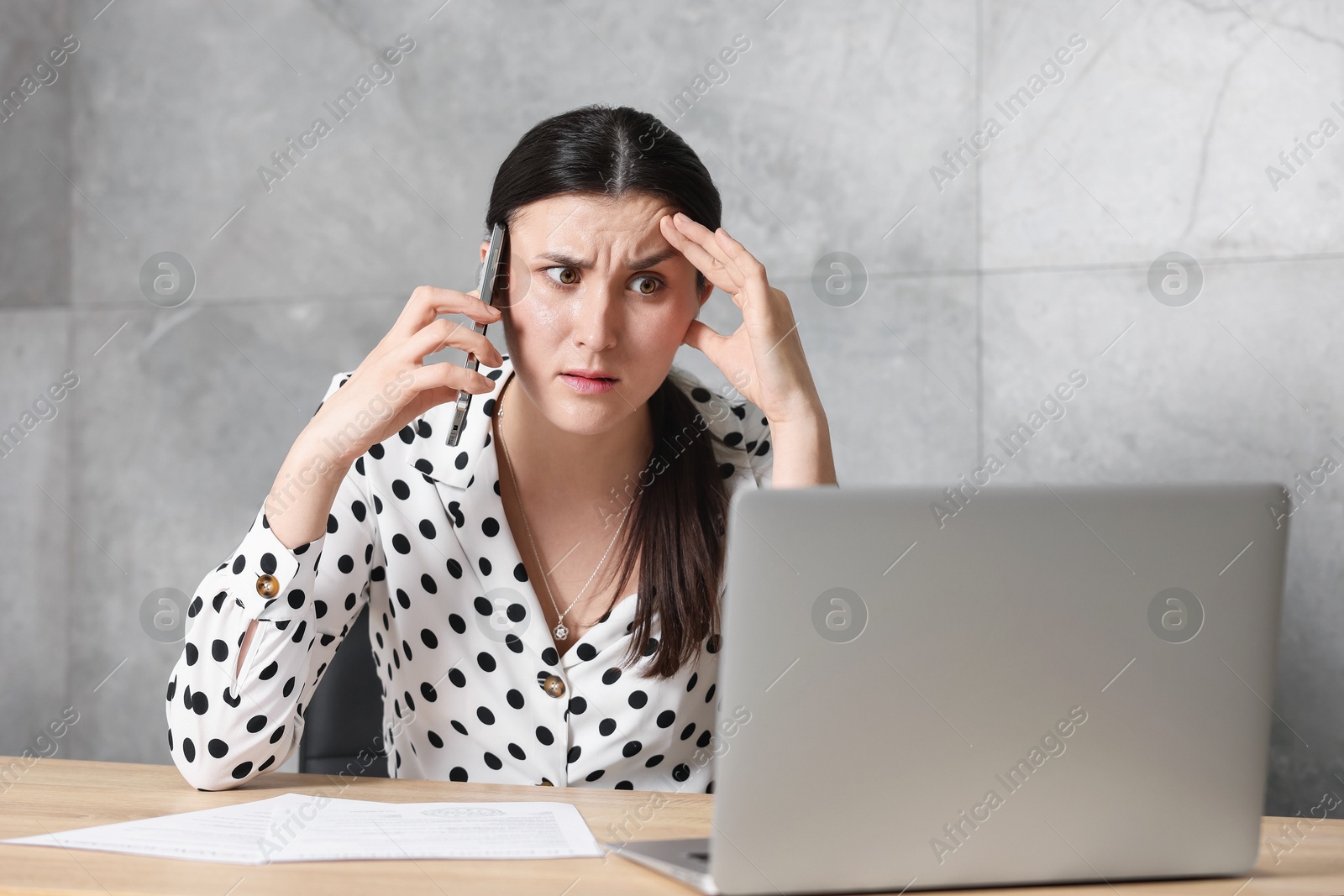 Photo of Young businesswoman with papers and laptop complaining to someone via phone at wooden table indoors