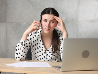 Photo of Young businesswoman with papers and laptop complaining to someone via phone at wooden table indoors