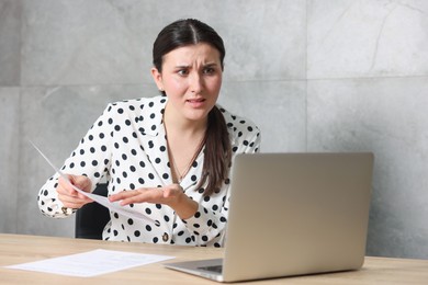 Photo of Young businesswoman with papers and laptop complaining to someone via phone at wooden table indoors