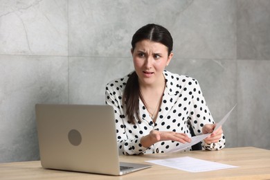 Photo of Young businesswoman with papers and laptop complaining to someone via phone at wooden table indoors