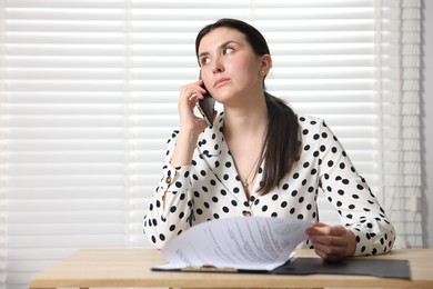 Photo of Young businesswoman with papers complaining to someone via phone at wooden table indoors