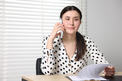 Photo of Young businesswoman with papers complaining to someone via phone at wooden table indoors