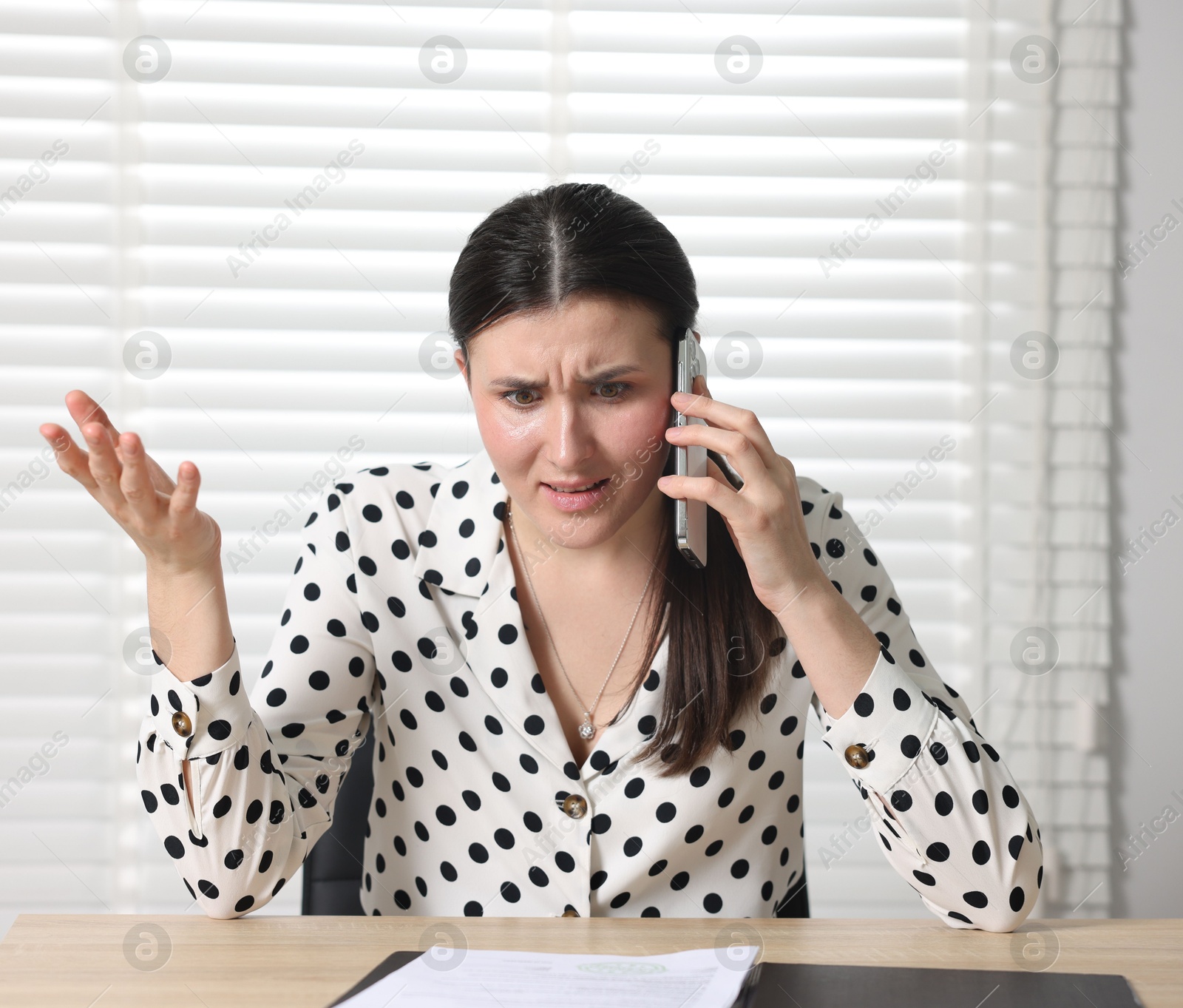 Photo of Young businesswoman with papers complaining to someone via phone at wooden table indoors
