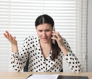 Photo of Young businesswoman with papers complaining to someone via phone at wooden table indoors