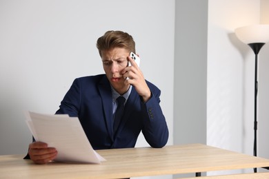 Photo of Young businessman with papers complaining to someone via phone at wooden table indoors